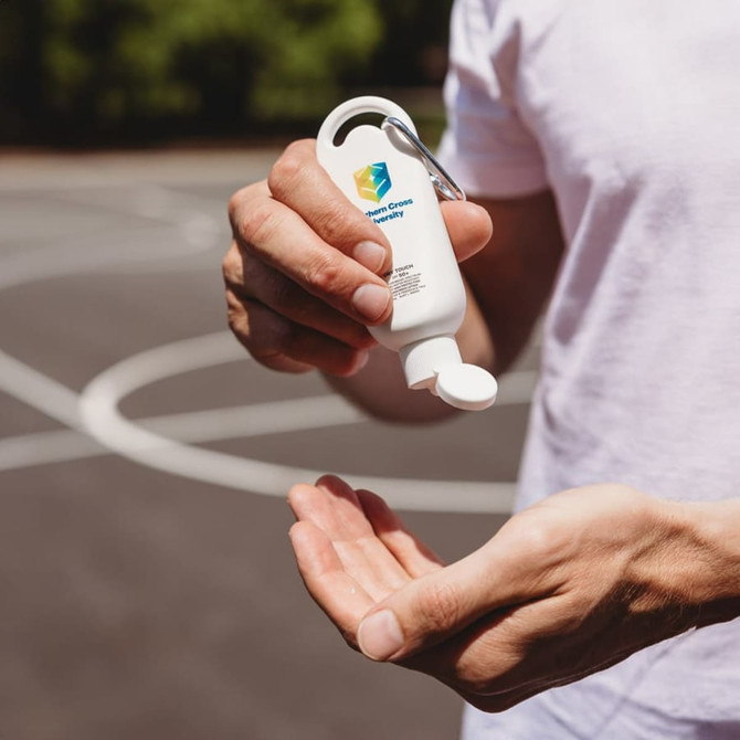 A white bottle of sunscreen with a flip cap held in a hand, featuring a logo and a carabiner attachment.