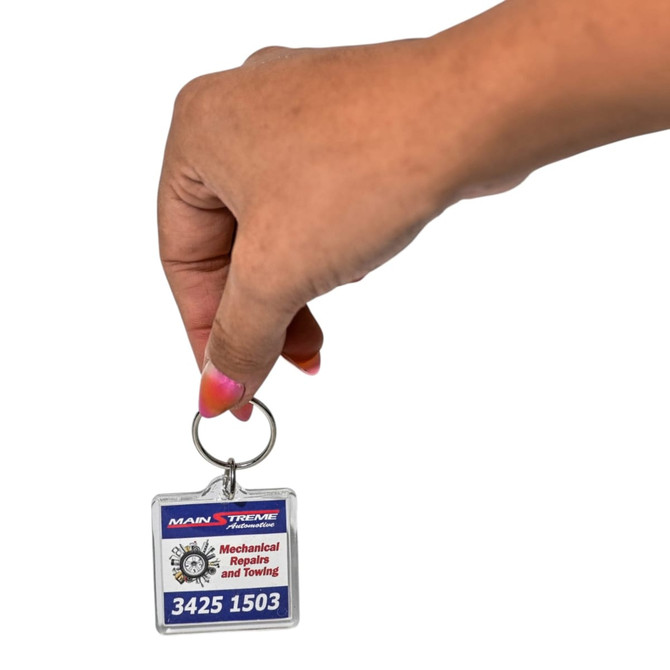 A hand holding a square acrylic keychain featuring a logo and text, predominantly red, blue, and silver.