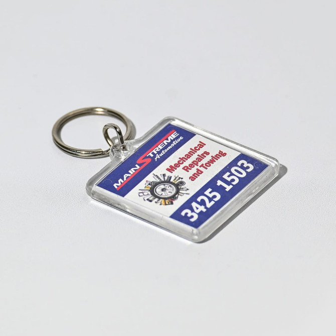 A square acrylic keychain with a clear border, featuring a logo and text in red and blue.