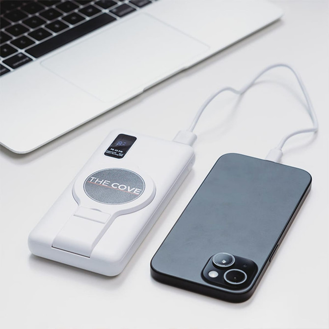 A white power bank with a circular logo, connected to a smartphone, placed on a light surface near a laptop.