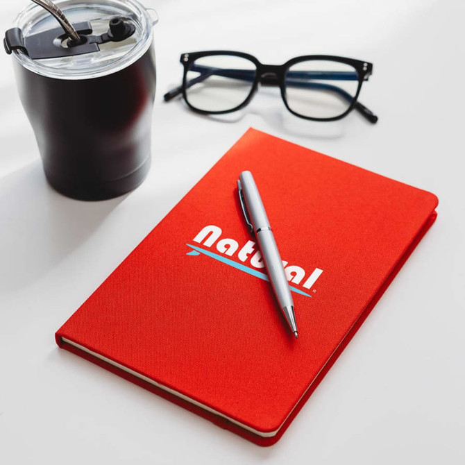 A red A5 notebook with a pen on top, beside a black drink bottle and glasses. The notebook features a logo.