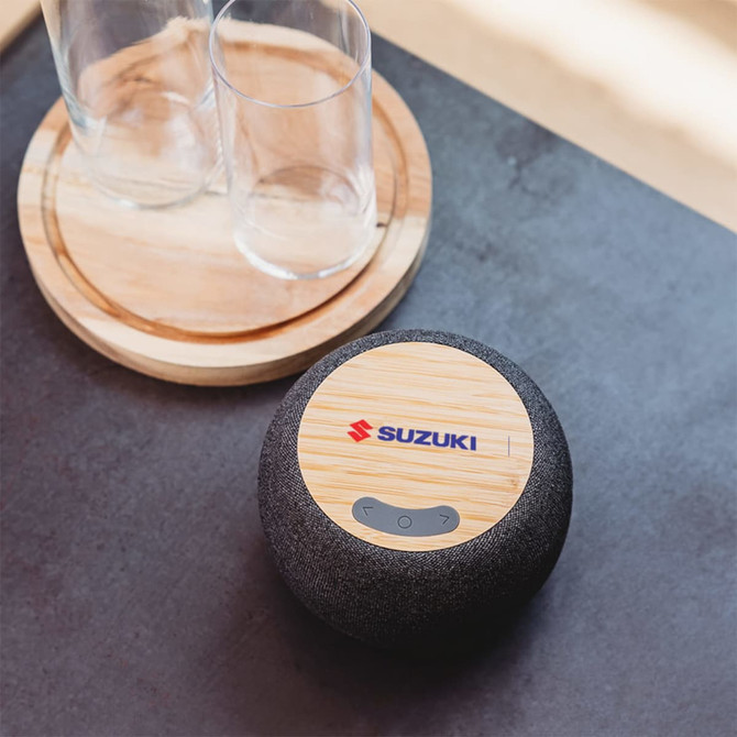 A round wireless speaker in black fabric with a bamboo surface on top, featuring a logo. Two clear glass cups are nearby.