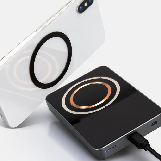A black wireless charging power bank with a circular charging area, beside a smartphone featuring a similar design element.