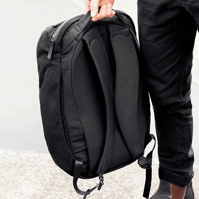 A black backpack with padded straps and a sleek design, held by a person. The bag features a branding logo.