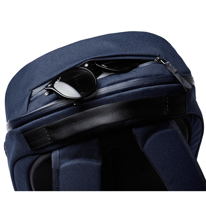 A navy Bellroy Via Backpack with a zippered compartment featuring sunglasses on top. The bag has a sleek, textured finish.
