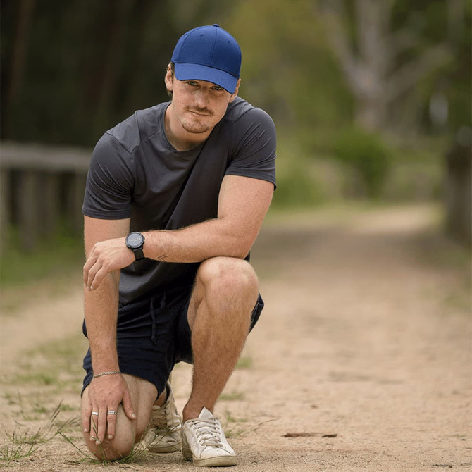 A man wearing a blue Falcon Flex Cap kneels on a sandy path, dressed in a grey shirt and shorts, with a watch on his wrist.