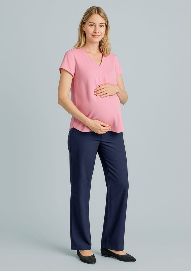 A pregnant woman wears a pink top and navy maternity pants, standing with one hand on her belly. The background is neutral.