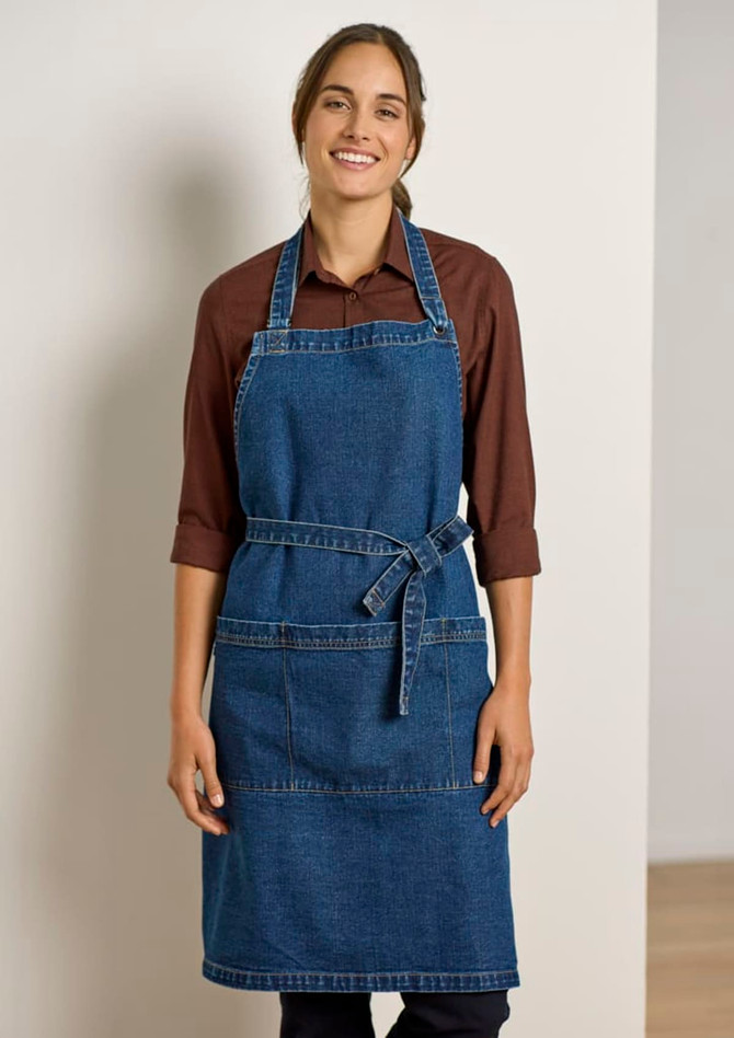 A denim apron in blue, featuring pockets and a waist tie, worn by a smiling woman. It has a logo.