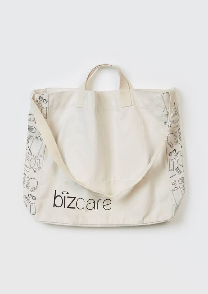 A beige unisex tote bag featuring a repeat pattern of various items and a logo. It has short handles and a long strap.