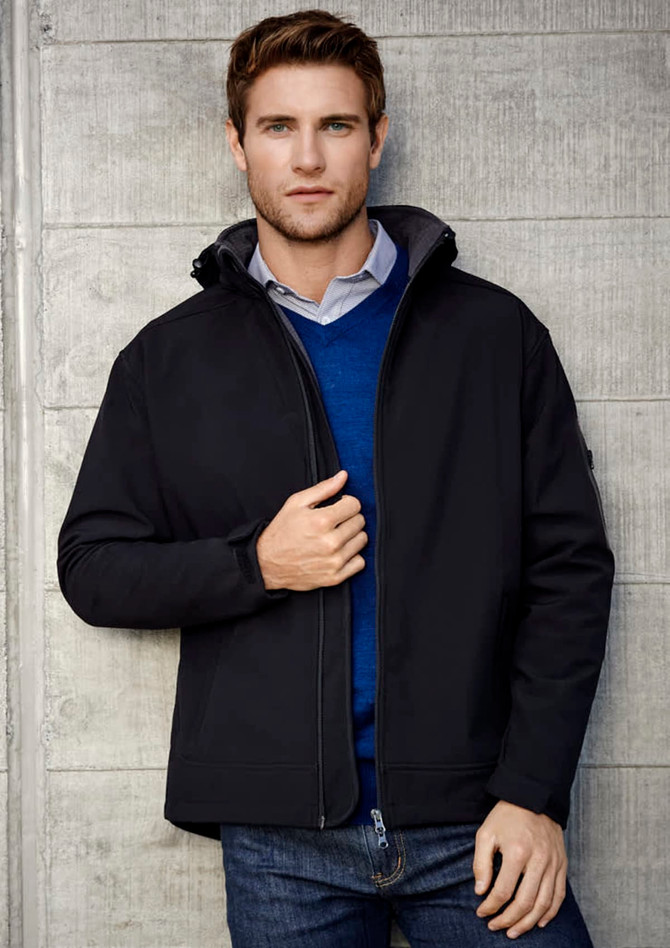 A black soft shell jacket is worn by a man against a concrete wall. The jacket has a hood and a logo on the sleeve.