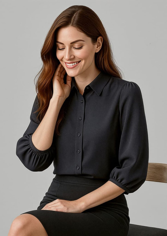 A woman in a black 3/4 sleeve blouse with a button front, sitting on a chair, smiling.