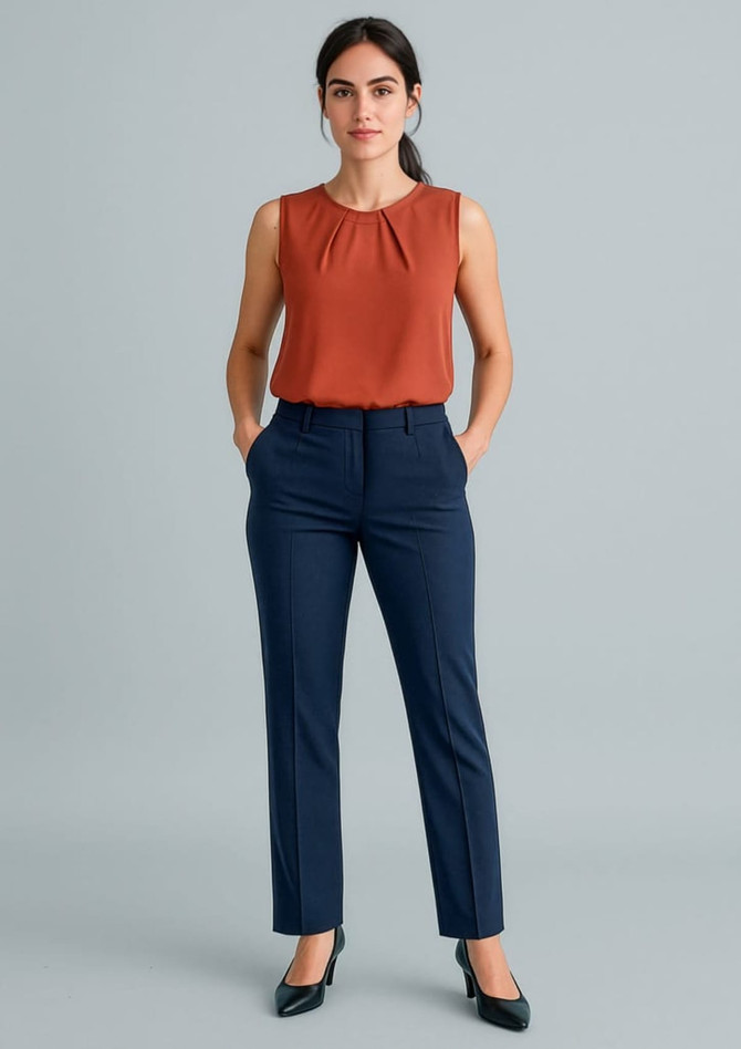 A woman in an orange sleeveless top and navy tapered leg adjustable waist pants, standing with hands in pockets.