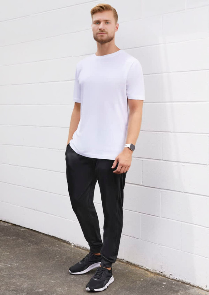 A man stands against a white wall wearing a plain white t-shirt and black track pants, with a watch and sneakers.