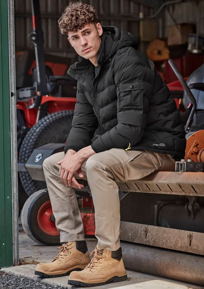 A man sits on a bench wearing khaki cargo pants and a black puffer jacket, with tan boots, in a rustic setting.