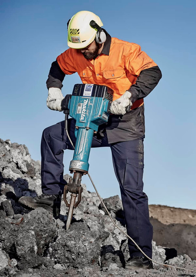 A man in safety gear operates a jackhammer on a rocky surface, wearing an orange shirt and navy blue cargo pants.