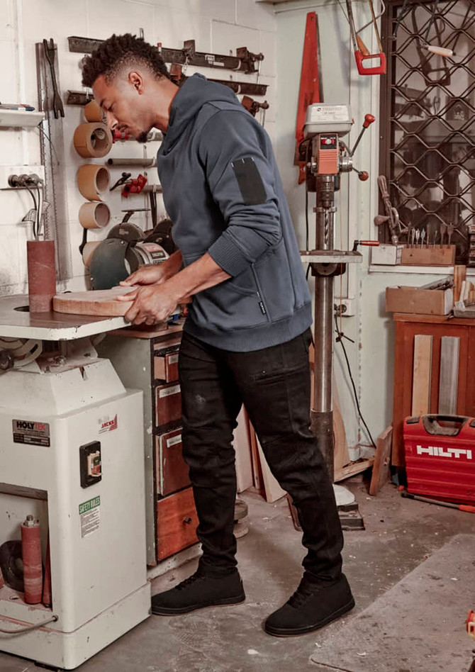 A man wearing a navy blue hoodie and black stretch jeans stands in a workshop, handling a piece of wood.