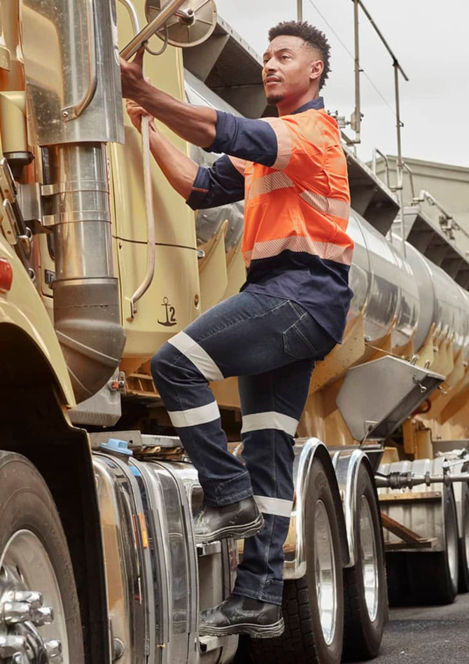 A man in an orange and navy work shirt climbs onto a truck, wearing dark blue stretch jeans with reflective stripes.