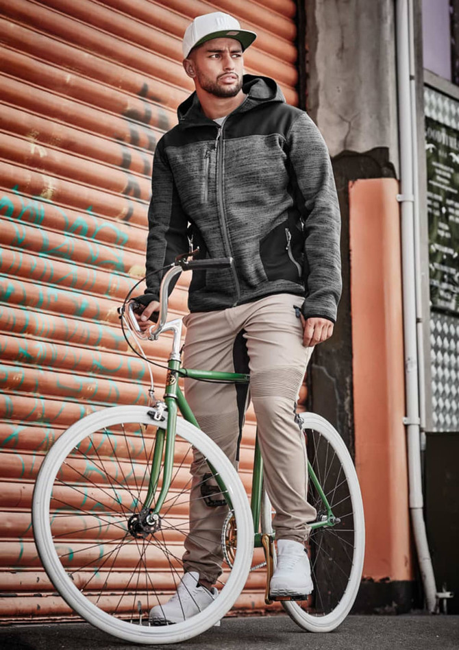 A man wearing a grey reinforced knit hoodie sits on a green bicycle against an orange corrugated backdrop.