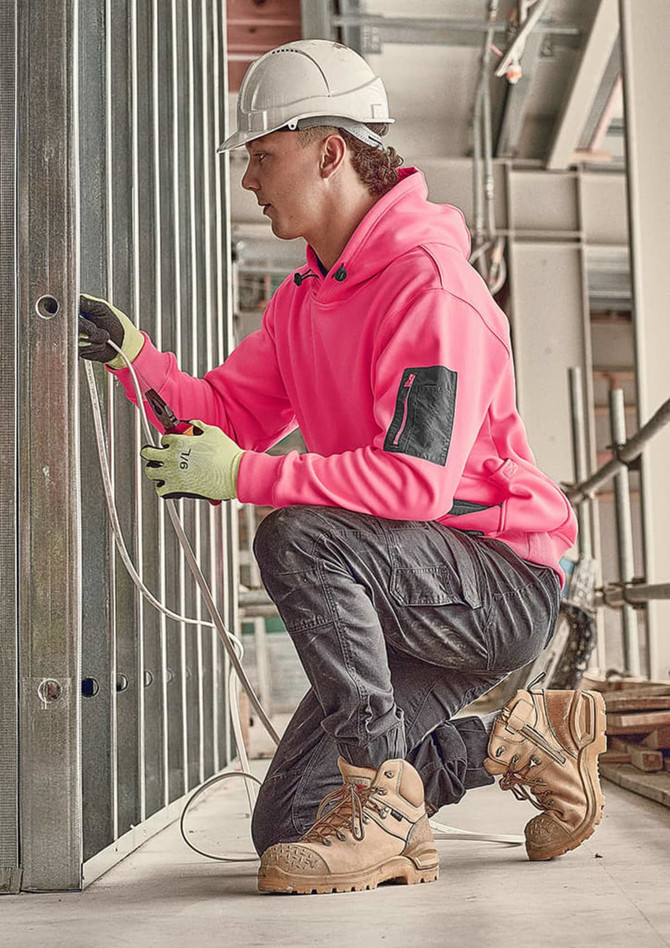A person in a bright pink water-resistant hoodie and grey work pants kneeling, working on electrical wires in a construction setting.