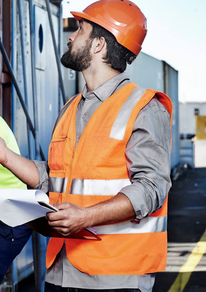 A man wearing an orange hi-vis zip vest with reflective stripes and a hard hat, holding documents at a construction site.