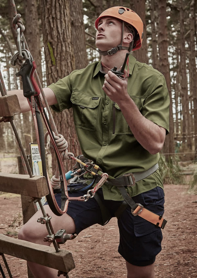 A man wearing a green short sleeve button-up shirt and blue shorts, equipped with climbing gear in a forest setting.