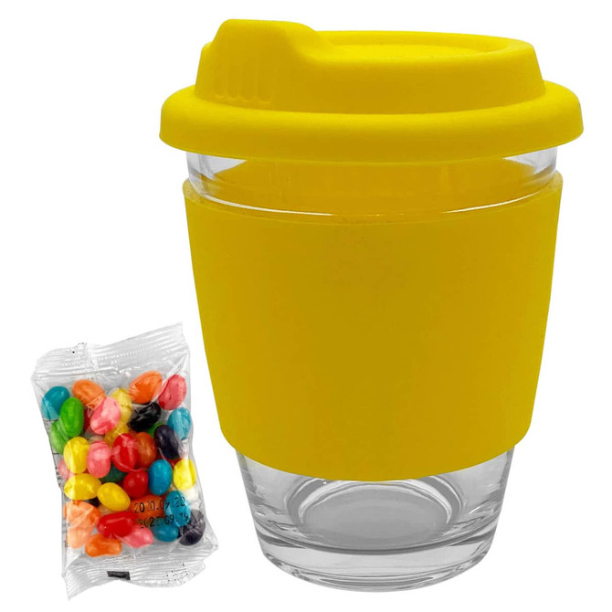 A clear glass coffee cup with a yellow silicone lid and sleeve, accompanied by a bag of colourful jelly beans.