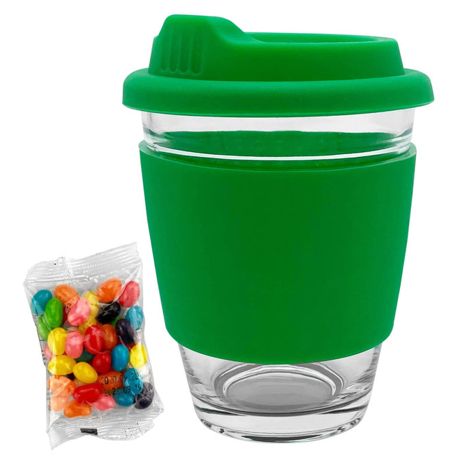 A clear glass coffee cup with a green silicone lid and sleeve, accompanied by a bag of colourful jelly beans.