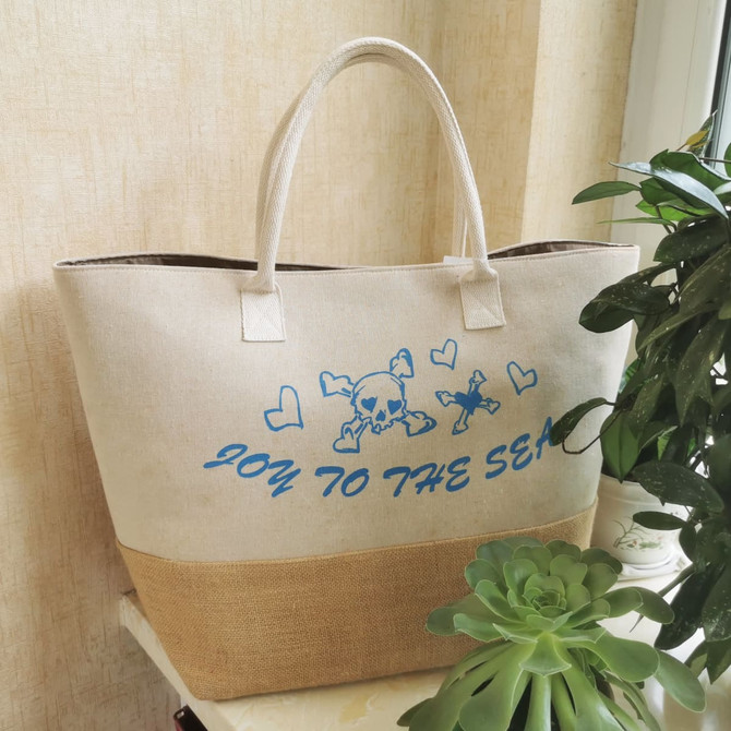 A large beige tote bag with a jute bottom and blue print design, featuring a logo and hearts.