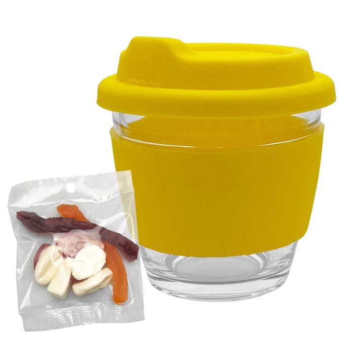 A clear glass coffee cup with a yellow silicone lid and sleeve, accompanied by a bag of mixed lollies.