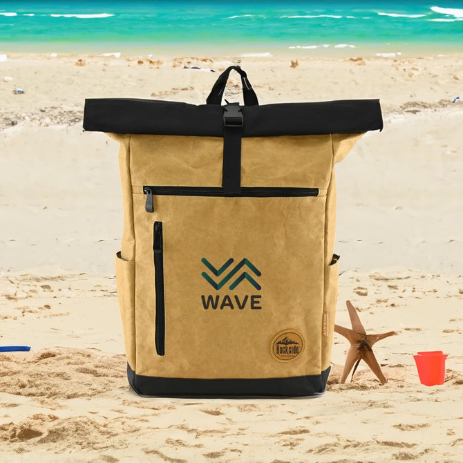 A khaki and black laptop backpack with a logo, placed on a sandy beach setting.
