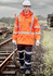 A man wearing an orange waterproof jacket and navy trousers walks along railway tracks, holding a tool. The jacket features reflective stripes.