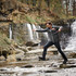A man in a grey rain jacket jumps over a rocky stream, with a waterfall in the background. The jacket features a logo.