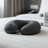 A grey neck pillow with a U-shape design rests on a neatly made bed. It has a small loop for attachment.