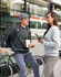 A grey crew fleece sweat worn by a woman and a dark grey version on a man, both casually styled outdoors with bicycles.
