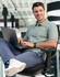 A men's cotton stretch polo in light green, featuring a logo, worn by a man seated at a desk with a laptop.