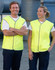 A hi-vis safety vest in bright yellow, worn by two smiling individuals. The vests feature a grey trim and have a logo.