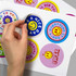 A hand peels a glossy sticker from a sheet featuring colourful circular designs with smiley faces and text.