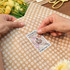 A person applies a decorative sticker to brown spotted wrapping paper, with yellow flowers in the background.