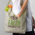 A cotton shoulder tote bag in light beige, featuring a logo and filled with lettuce and a drink bottle.