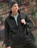 A men's sustainable softshell jacket in black, featuring a zip front and a logo, worn with a beanie and backpack.