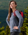 A woman wearing a long-sleeve grey tee shirt and a red and black backpack stands outdoors against a forest backdrop.