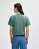 A green women's boxy t-shirt, viewed from the back, paired with light-coloured jeans. It features a branding logo.