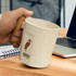 A coffee mug in a light, speckled cream colour with a wooden handle, featuring a logo. Steam rises from the mug.