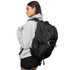 A black backpack with multiple compartments is worn on one shoulder by a person in a grey hoodie.