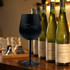 A black stainless steel wine glass beside several green wine bottles on a wooden surface. The glass has a branding logo.