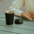 A double-walled black coffee cup with a lid, placed on a green table alongside a person's hand.
