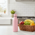A drink bottle in pink, featuring a sleek design, sits on a kitchen counter beside a basket of fresh fruit.