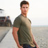 A model wearing an olive green Men's CVC Crew T-shirt stands on a beach pathway, with a blurred background of buildings.