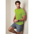 A man is wearing a bright green t-shirt and grey shorts, leaning against a wall. The shirt has a logo.