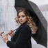 A black umbrella with a curved wooden handle, held by a person with long, wavy hair. The umbrella has a logo.